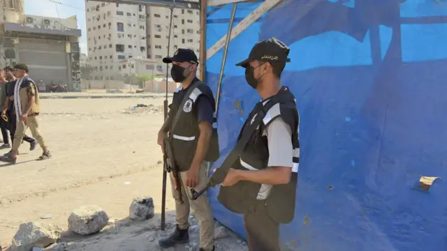 Two gunmen hold their guns in front of a blue tarpaulin
