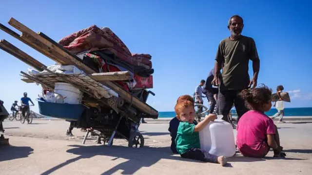 A toddler with red hair holds an empty container for water, his father and siblings are behind him with their cart