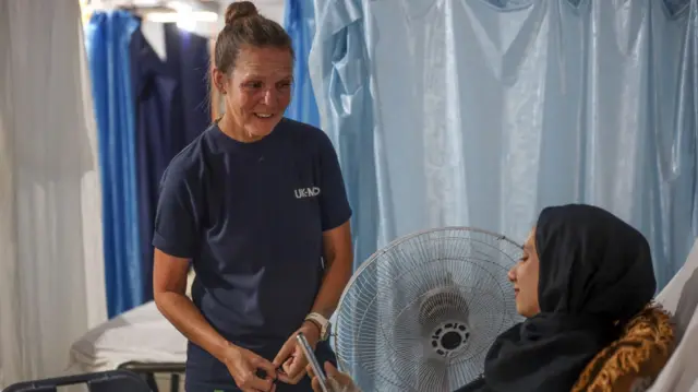 Paula Tobin stands at the bedside of a patient in Gaza who is looking at her phone while conversing. Paula has her hair up in a bun and is wearing a T-shirt with the UK-Med logo on it. The patient is a woman wearing a black headscarf.
