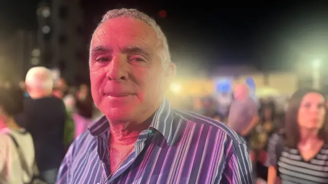 Elderly man in striped shirt looks into the camera as he attends a rally in Hostages Square, Tel Aviv. Behind him are several other people, partially blurred