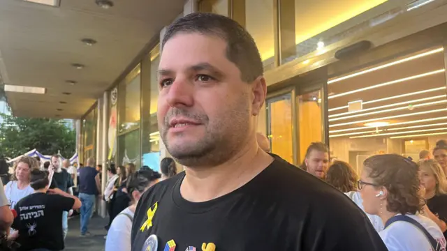 Omri Lifshitz, a middle aged man in a short-sleeved black shirt with a yellow ribbon pin to the front, stands among the crowd on the sidelines of Hostages Square