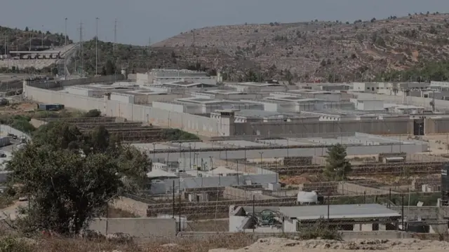 A wide-shot of a grey and white prison building surrounded by fences. A tree is in the foreground of the picture and hills are visible behind the prison.