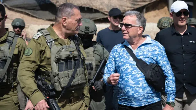 Steve Witkoff in a blue camouflage print shirt with a white t-shirt underneath, he is walking with military personnel.