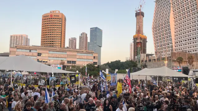 Large crowd assemble in Hostage Square in Tel Aviv in the late afternoon, the skyline of the city in the background. Some people in the far right of the frame are holding two US flags, while others hold placards with pictures of the hostages or Israeli flags