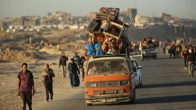 A battered, orange Volkswagen van travelling on a main road is piled high with goods on its roof, including chairs, blankets and suitcases. People are also walking along the sides of the road.