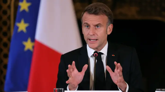 Close up of Emanuel Macron with his hands raised as he sits in a black suit. Behind him are a French and EU flag