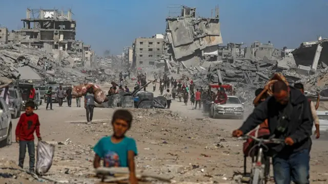 A group of people walk on road, rubble and collapsed buildings behind them.