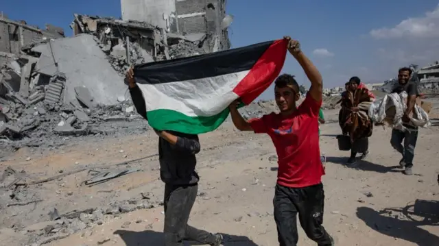 Two men carry a Palestinian flag above their heads as they walk towards their homes in the Khan Younis area. People carrying belongings walk behind them. Destroyed buildings are in the backdrop.