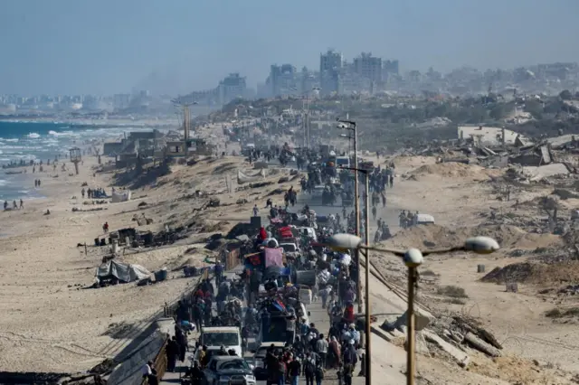 People walking on foot are interspersed with vehicles piled high with goods making their way up to the north of Gaza along a narrow, coastal road. High-rise buildings are in the distance, while the beach and sea is to the left of the road.