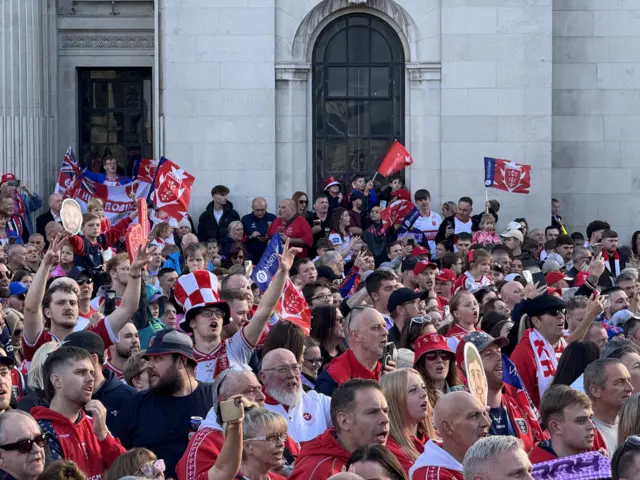 A crowd of people wearing red hats, scarves and waving flags are signing