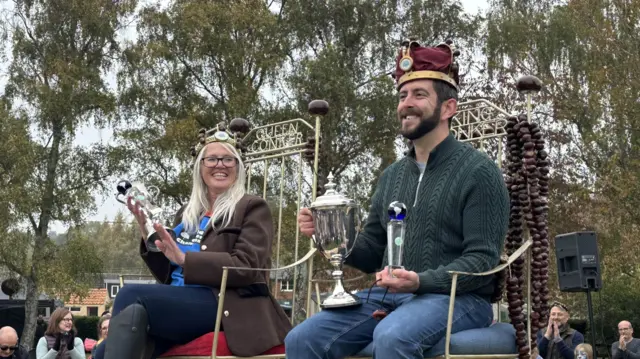 Two people sat on golden thrones and were holding trophies. They both were wearing crowns.