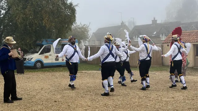 People were dressed in morris dancing clothing, wearing white shirts and black shorts with white socks. They were dancing on gravel.