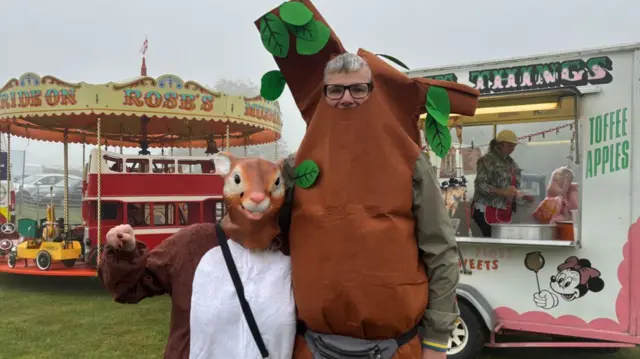 Two people in fancy dress - one as a squirrel and another as a tree - stand next to each other in front of a candy floss stand and a merry-go-round. The sky is foggy and grey