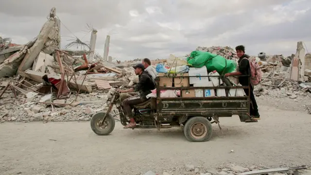 A man drives a motorised tricycle with a flatbed attached to it carrying several boxes. There are two passengers, one at the front and one at the back. It's drving through a destroyed city in Gaza