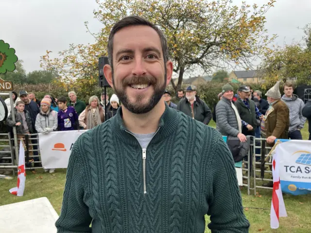 Man smiling as he looks to camera. He's wearing a green knitted jumper.