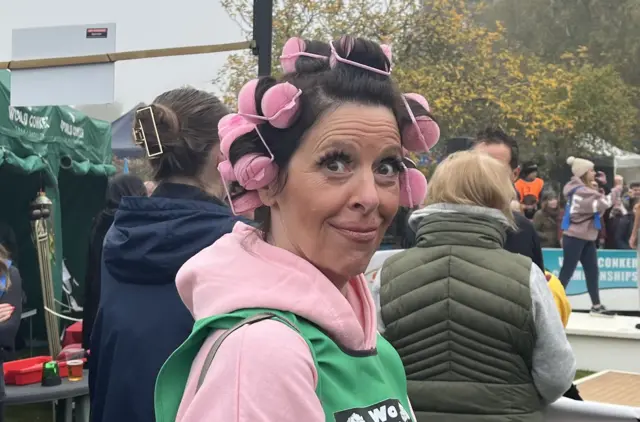 Emma has her hair in pink rollers which are on her head. She is wearing a pink hooding and is standing in front of an arena where people are playing Conkers.