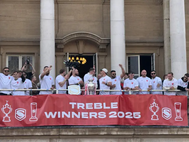 A team of players wearing white T-shirts are lined up on the Hull City Hall balcony. There is a banner in front of them that says Treble winners 2025.