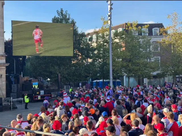 A crowd of fans wearing red hats, scarves and shirts are gathered in a city centre, looking up at a big screen which is playing a rugby match between Hull KR and Wigan.