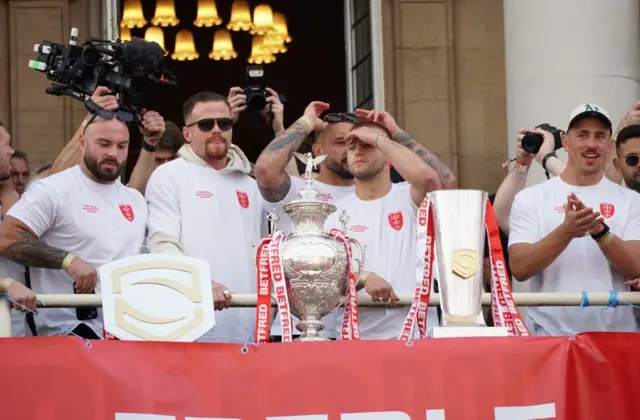 Five Hull players are standing on the balcony of Hull City Hall and looking out towards the crowd. There are three silver tropohies in front of them