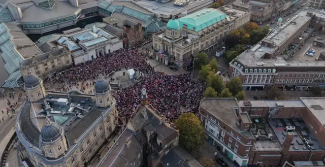 An aerial view of Hull City Centre as thousands of people wearing red gather in a city square. They are surrounded by a large shopping centre and tall buildings.