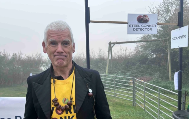 A man with grey hair is wearing a yellow tshirt, black jacket and has a string of conkers around his neck. He is standing in a field in front of two white signs that say 'steel conker detector' and 'scanner'