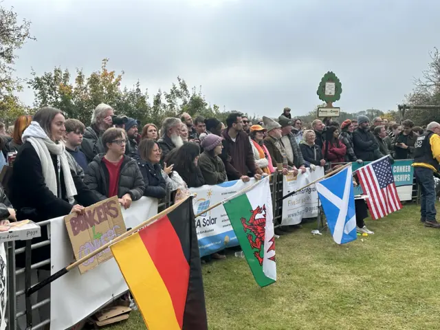 A crowd of people are standing behind a barrier which has sponsor advertisement attached to it.