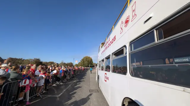 A white bus pictured with a line of fans standing next to it.
