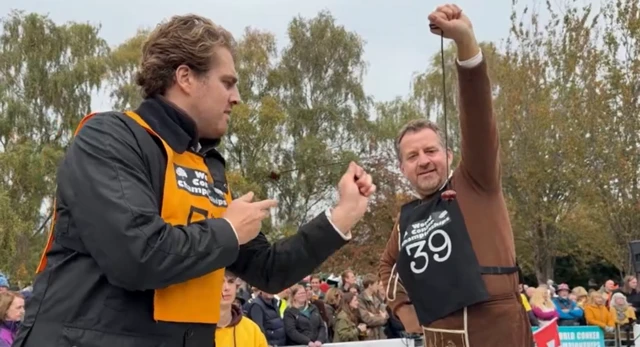 A man in a brown jumper holds up a conker on a black string while another man wearing black aims for it with his.