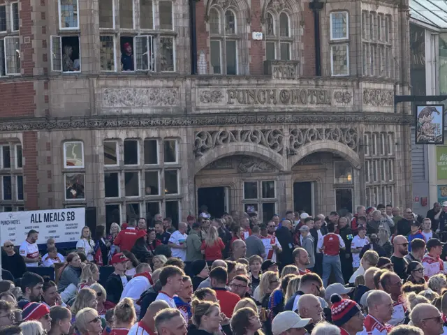 A crowd of people outside a pub. They are standing with drinks in their hands and looking towards the main city hall