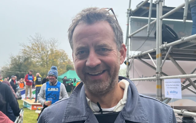 A smiling man looks to the camera. He has spectacles on the top of his head and is stood in front of scaffolding.