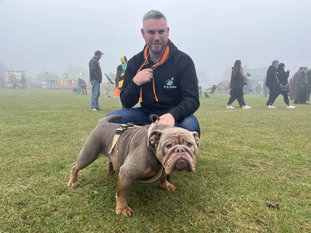 Tayah is a brown small Old English Bulldog standing in front of her owner on a field. Lee is wearing a black jumper.