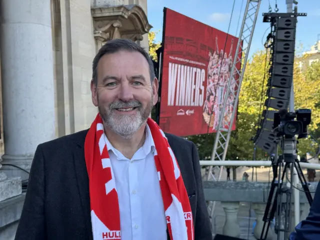 A man with short dark hair is wearing a black suit coat and white shirt, with a red scarf around his neck. He is smiling to the camera.