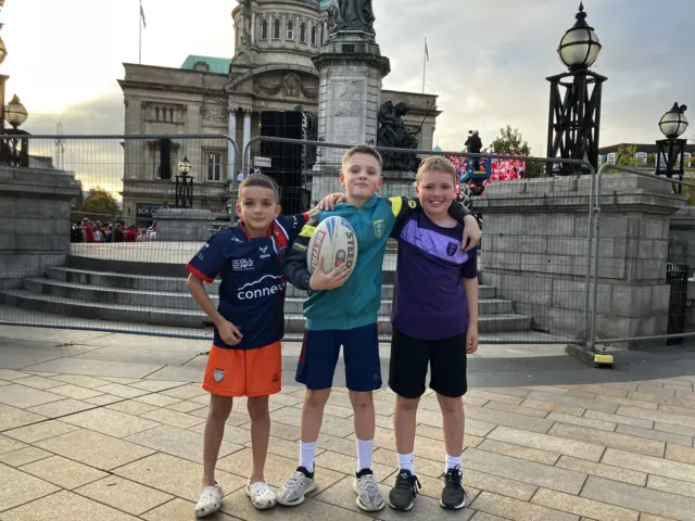 Three young boys standing with their arms around each other. The boy in the middle is holding a rugby ball