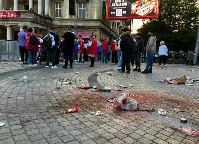 A shot of rubbish on the floor with a carrier bags and bottles in the foreground and people standing around in the background.