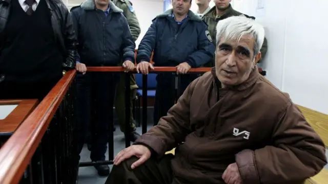 Ahmed Sadaat looks at the camera while he sits in an area enclosed by a wooden railing at a military court in Israel with policemen flanked around him on the other side of the barrier. The picture was taken in 2008.