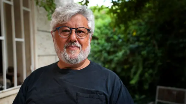 A middle aged man with white and grey hair wearing a short sleeved blue t-shirt looks into the camera while standing in a residential garden