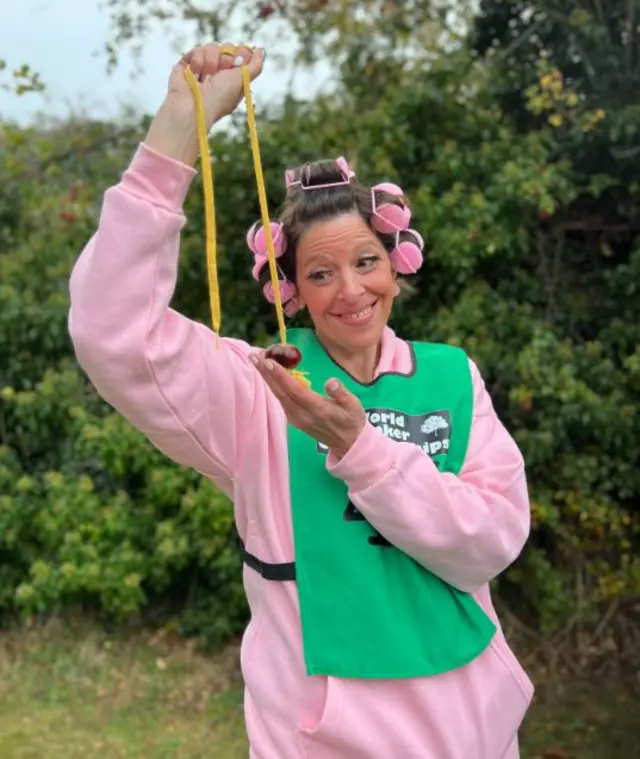 Lady with pink top on and green bib smiles at a conker on a string. She has pink rollers in her hair and is stood in front of a green bush.
