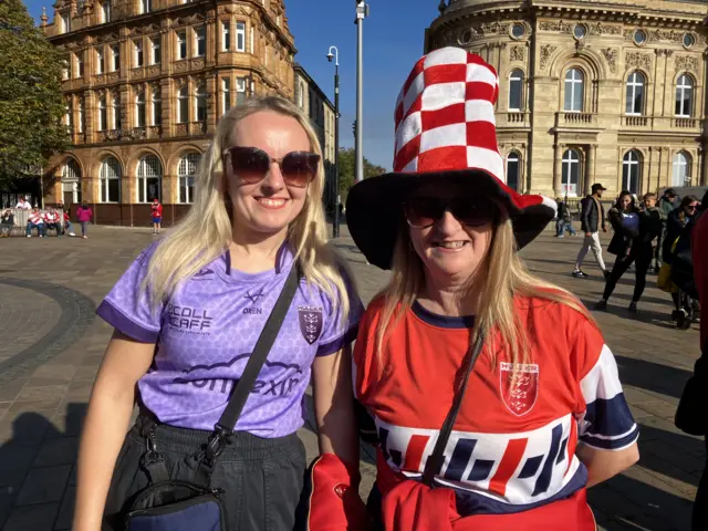 Two women standing in the middle of a city centre square. One has a purple rugby shirt with blond hair and sunglasses while another has a red shir and is wearing a red and white top hat.