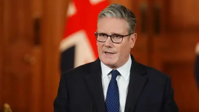 Sir Keir Starmer speaks in a wood-walled room in front of two union jack flags. He is wearing a black suit jacket, a blue tie with white polka dots, and a white shirt. He has a serious expression.