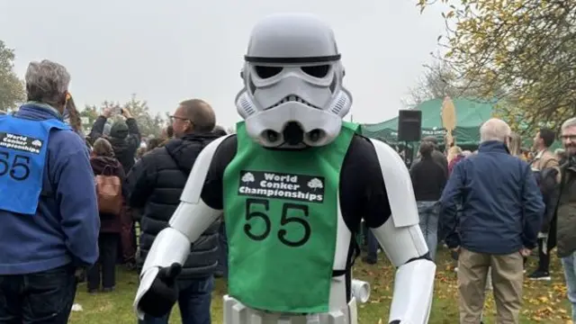 Stormtrooper standing in a field with a crowd of people facing the other way behind him. On his costumer is a green bib with World Conker Championships written on it and the number 55