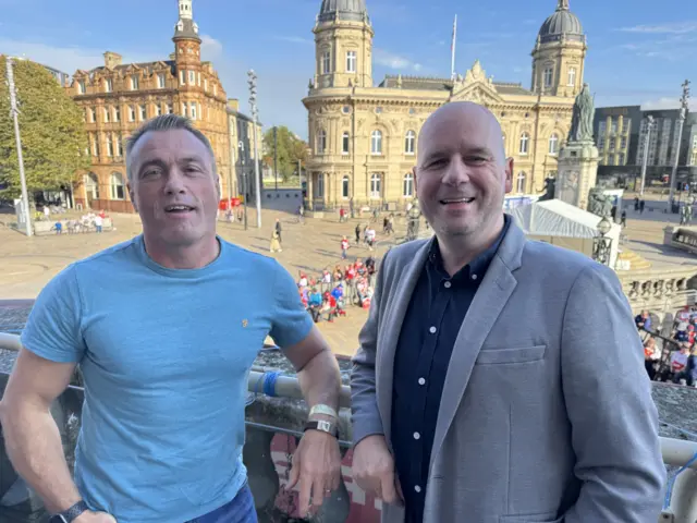 A man with a tight-fitting blue t-shirt on with short light brown hair is standing on a city hall balcony next to a man with a bald head, grey suit jacket and blue shirt.