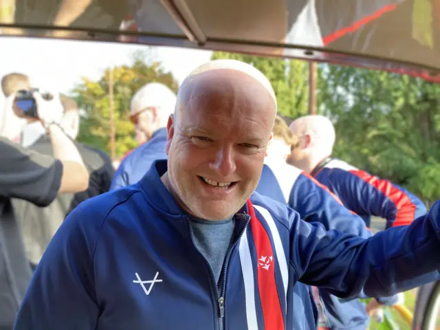 A man with a bald head is smiling to the camera and is wearing a blue tracksuit with red and white stripes down one side