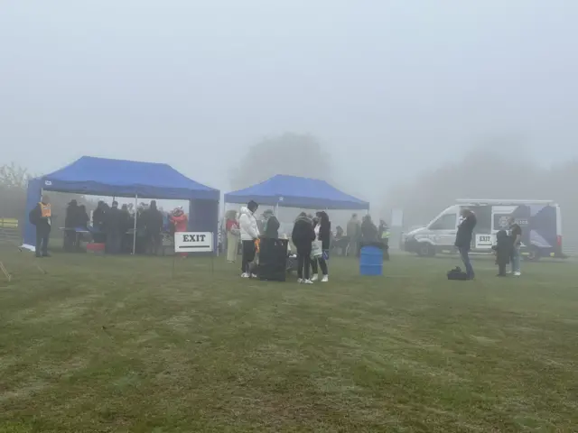 Two blue tents were situated on a field to the left of the image. People were walking through the tents and standing on the field.