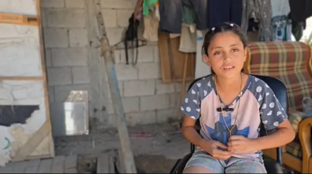 Tala sits on a chair wearing a patterned T-shirt and shorts. Behind her is an armchair and clothes which appear to be hanging up drying.