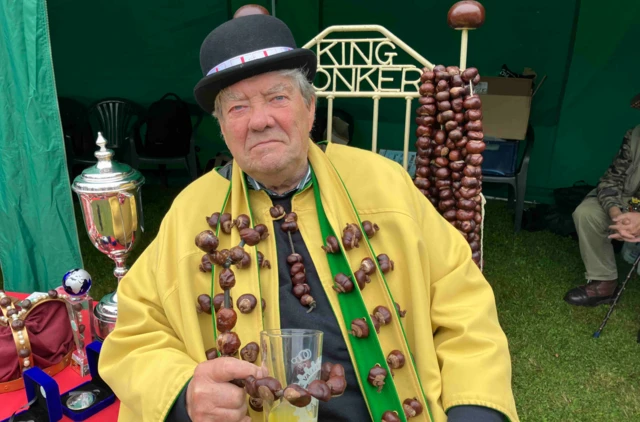 A man in a yellow jacked and black bowler hat with strings of conkers around his neck