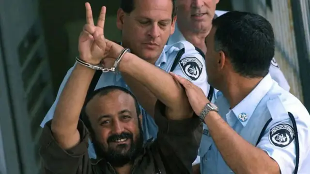 Marwan Barghouti  is seen in handcuffs as he flashes a peace sign with his fingers. Officers hold his arms as they escort him - he is smiling