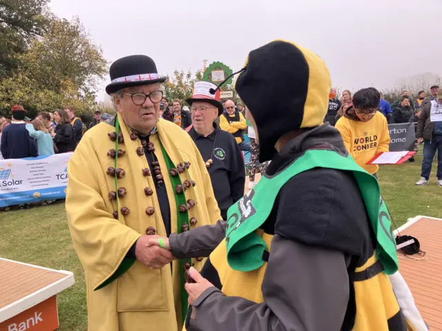 Two people are shaking hands standing in the middle of an arena. The man on the left is wearing a yellow coat with a conker necklace, and the woman on the left is a wearing a bumble bee costume.