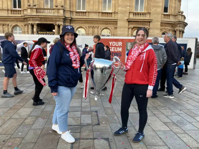 A young girl with a red jumper and a woman with a blue hoodie and hat are standing next to each other and holding an inflatable trophy.