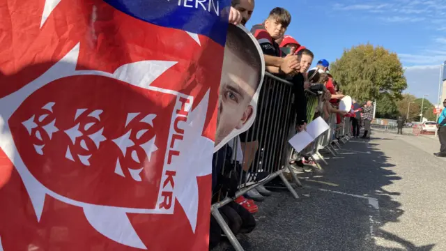 A line of fans, with red flags, standing behind metal security barriers in the car park of a rugby ground.