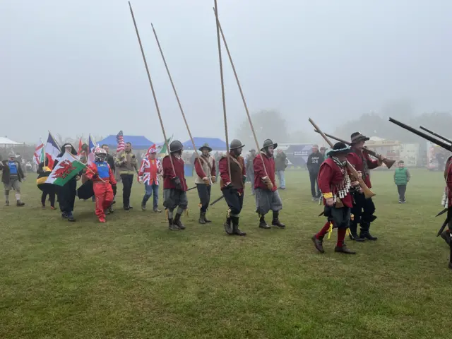 People dressed in different costumes were walking on a field. Some of them were carrying long wooden sticks in the air.